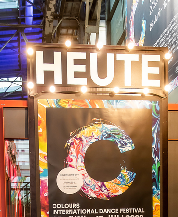 Es ist eine große Pinnwand zu sehen. Die Pinnwand steht im Theaterhaus. Auf der Pinnwand ist das Zeichen vom COLOURS Tanzfestival.