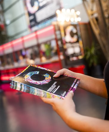 Im Theaterhaus Foyer. Das Foyer ist prächtig im Look des COLOURS International Dance Festivals. Zwei Hände halten einen Stapel Programmflyer.